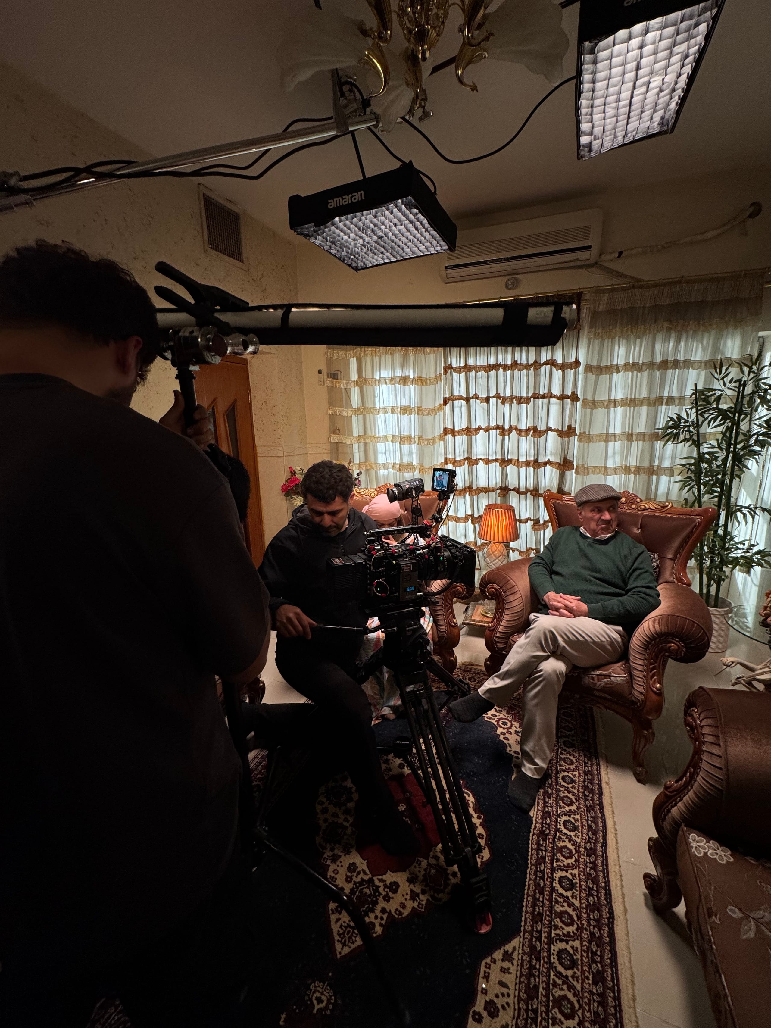 Фотография, сделанная на съёмочной площадке, в полутёмной комнате с фактурными кремовыми стенами. Мужчина с седыми усами, в серой клетчатой кепке, тёмно-зелёном свитере и бежевых брюках сидит, закинув ногу на ногу и сцепив руки, в резном деревянном кресле, обитом блестящей коричневой тканью. Рядом с ним горит настольная лампа с оранжевым плиссированным абажуром; за его спиной светятся полупрозрачные шторы с оборками. Слева за камерой на штативе с небольшим монитором сверху сидит мужчина в чёрной толстовке с капюшоном; человек в розовом головном уборе частично скрыт за камерой. На переднем плане мужчина, которого видно со спины, держит вертикальную штангу; на её верхнем конце зажим удерживает длинную светлую трубку. Под потолком, рядом с люстрой золотистого цвета, висят две включённые световые панели с сотами, на одной из них надпись «amaran».