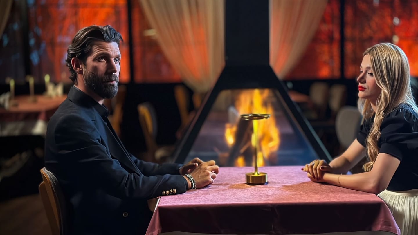 A bearded man in a dark suit and a blonde woman in a black top and cream pleated skirt sit across from each other at a table under a dusty-rose cloth, the camera looking down its length. Each rests clasped hands on the cloth, which is otherwise bare but for a slim gold object with a round base and a flat disc on top; directly beyond it a large flame burns inside a tall angular dark-framed enclosure. He is turned toward the camera, she is in profile with her gaze lowered. Out of focus around them, a dim dining room: a bare round wooden table holding tall pale candles in candlesticks, cream pleated curtains, orange-lit panels.